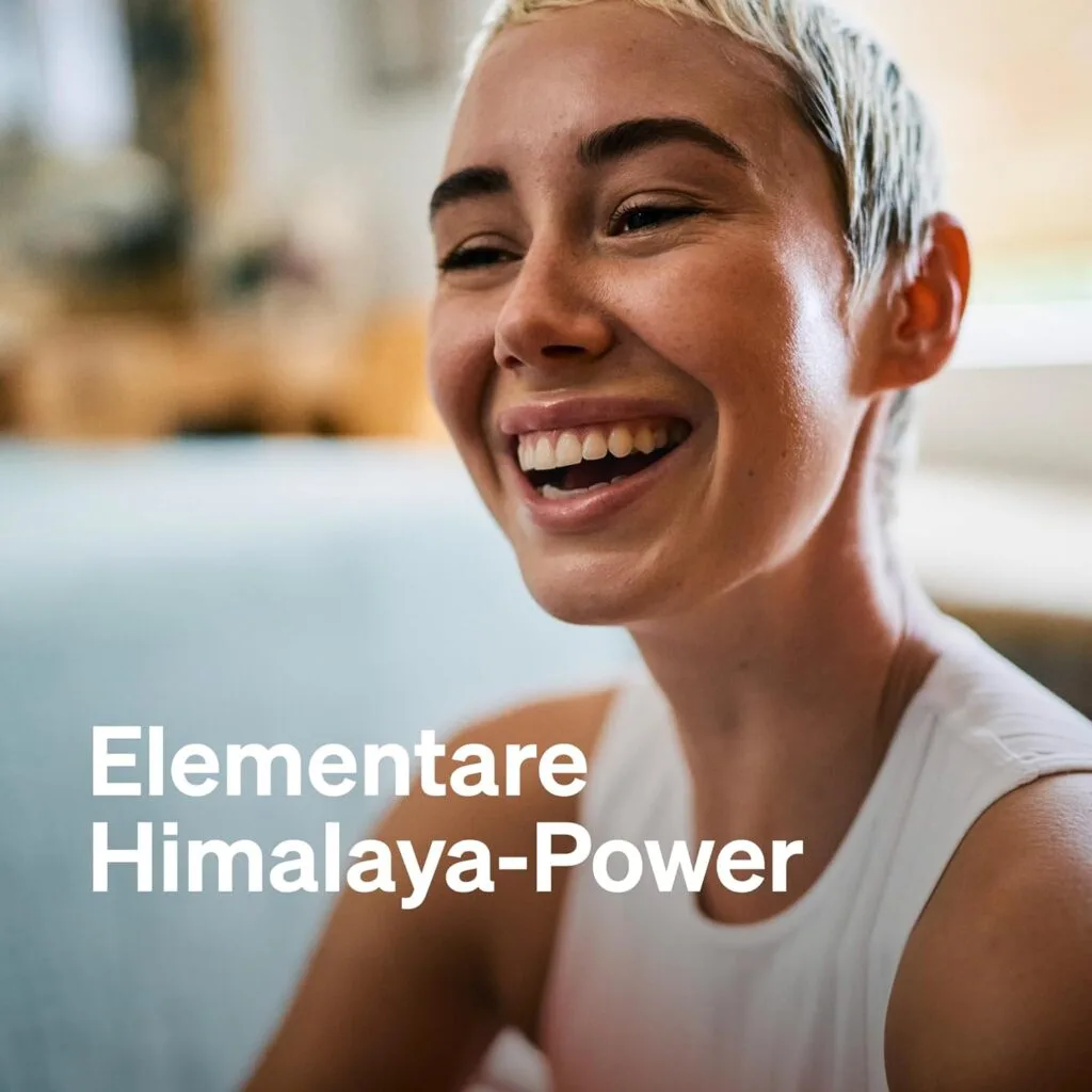 Une personne aux cheveux blonds courts sourit à l'intérieur. Le texte blanc sur l'image indique "Elementare Himalaya-Power", mettant en évidence Natural Elements Shilajit 1300mg - 120 Gélules Hautement Dosées, Mumijo de l'Himalaya Original, vegan & lab-tested.