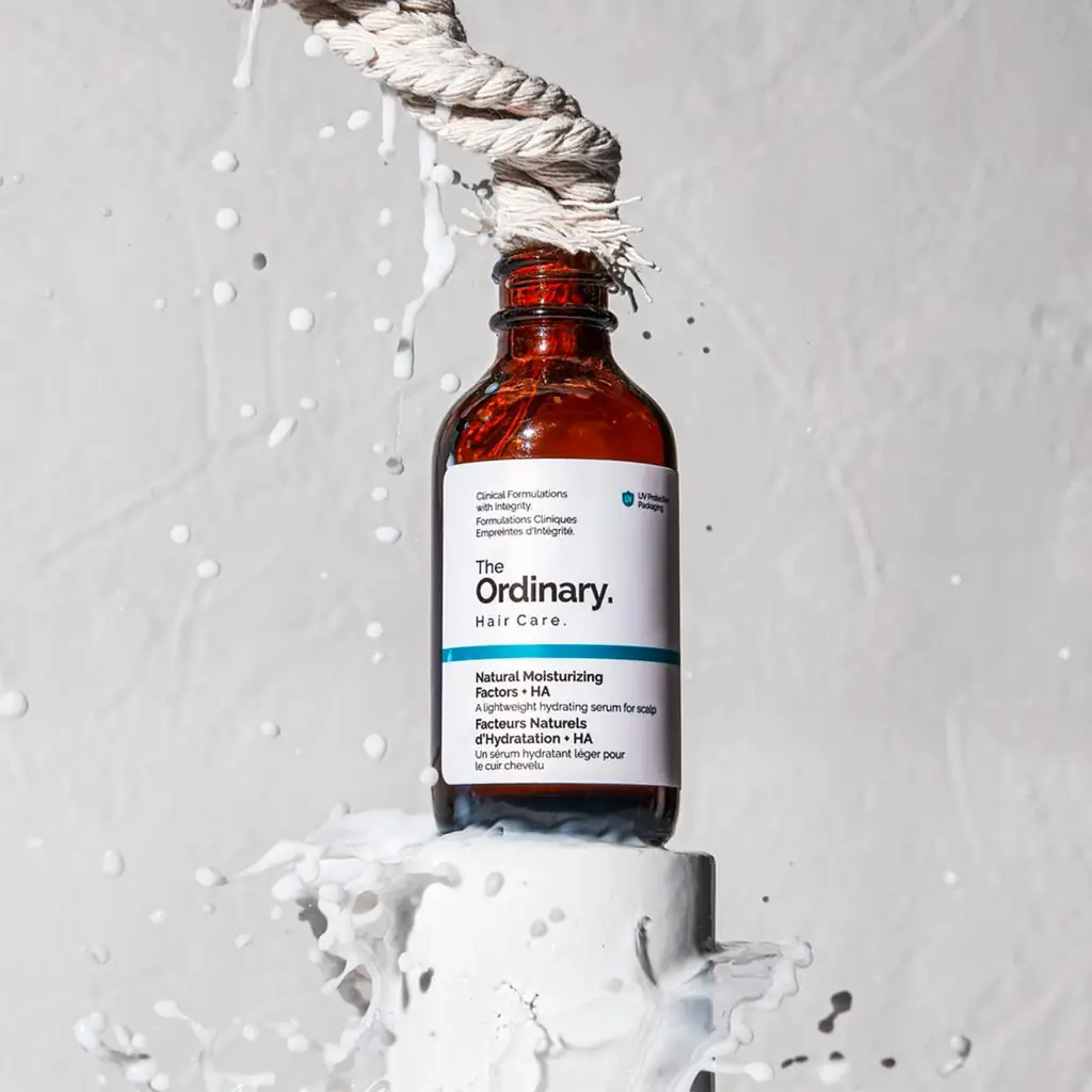 Un flacon en verre brun de The Ordinary NMF + HA Sérum Cuir Chevelu - Hydratation & Squalane (60 ml) pend au bout d'une corde épaisse, le liquide éclaboussant à sa base sur un fond gris.
